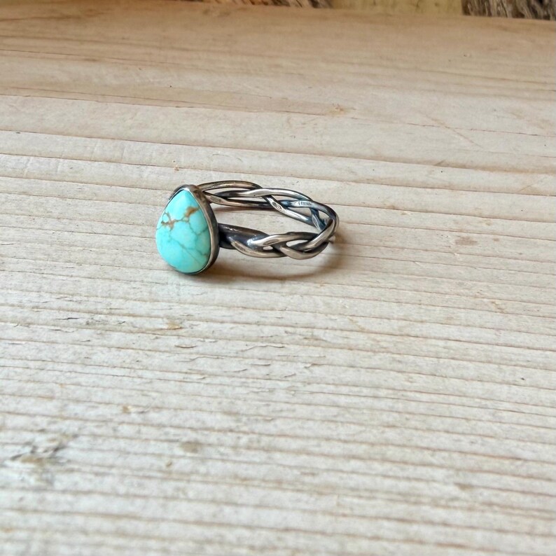 Puede incluir: Un anillo con una piedra turquesa en forma de l&aacute;grima con vetas marrones, engastada en una banda de metal oscuro con un dise&ntilde;o trenzado y retorcido. La piedra est&aacute; situada en la parte superior de la banda, que tiene un acabado oxidado y texturizado.