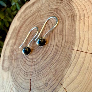 May include: A pair of silver drop earrings featuring round, polished black stones set in scalloped metal bezels. The earrings have long, curved silver ear wires that extend upward from the stone settings, creating a simple and clean silhouette.