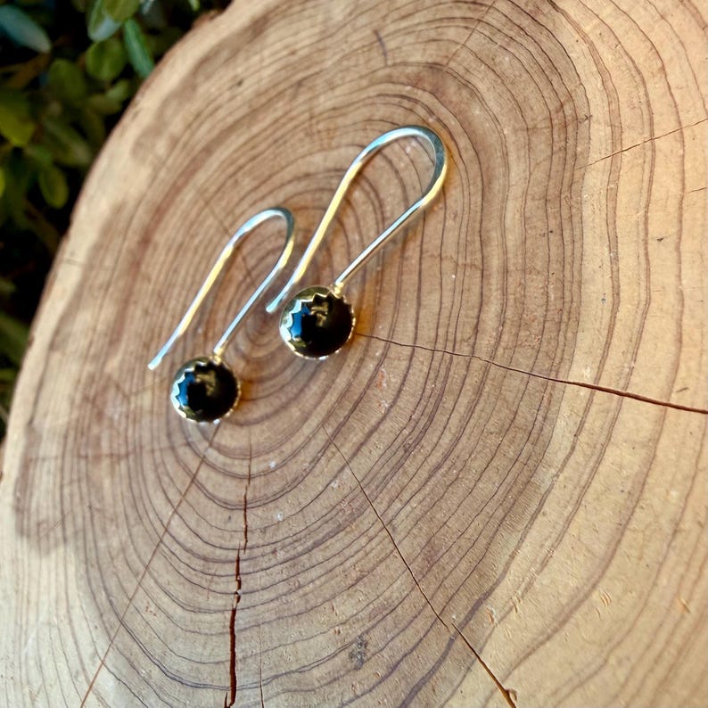 May include: A pair of silver drop earrings featuring round, polished black stones set in scalloped metal bezels. The earrings have long, curved silver ear wires that extend upward from the stone settings, creating a simple and clean silhouette.