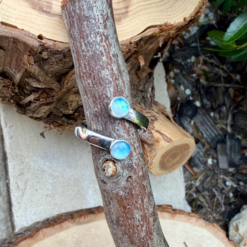 May include: A silver ring featuring two circular, light blue iridescent stones. The ring is displayed on a brown wooden branch, highlighting the jewellery's design and colour against the natural wood grain.