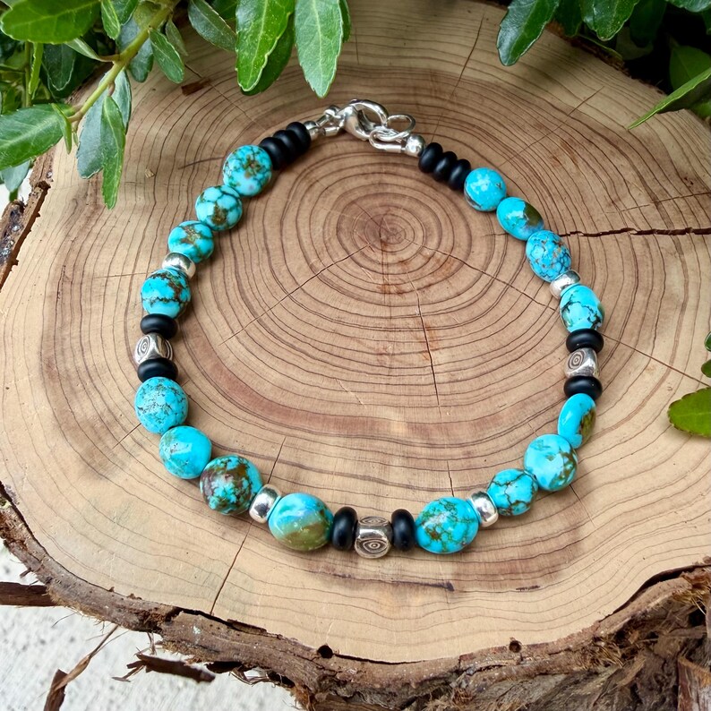 May include: A turquoise beaded bracelet with silver accents and a silver clasp. The bracelet features round, turquoise beads interspersed with black and silver beads. The bracelet is displayed on a wooden surface.
