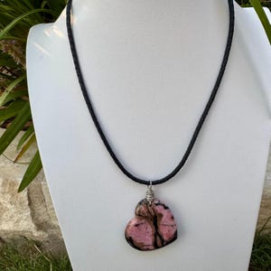 May include: A heart-shaped pendant necklace featuring a pink and brown stone. The pendant is suspended from a black braided cord. The necklace is displayed on a white mannequin.