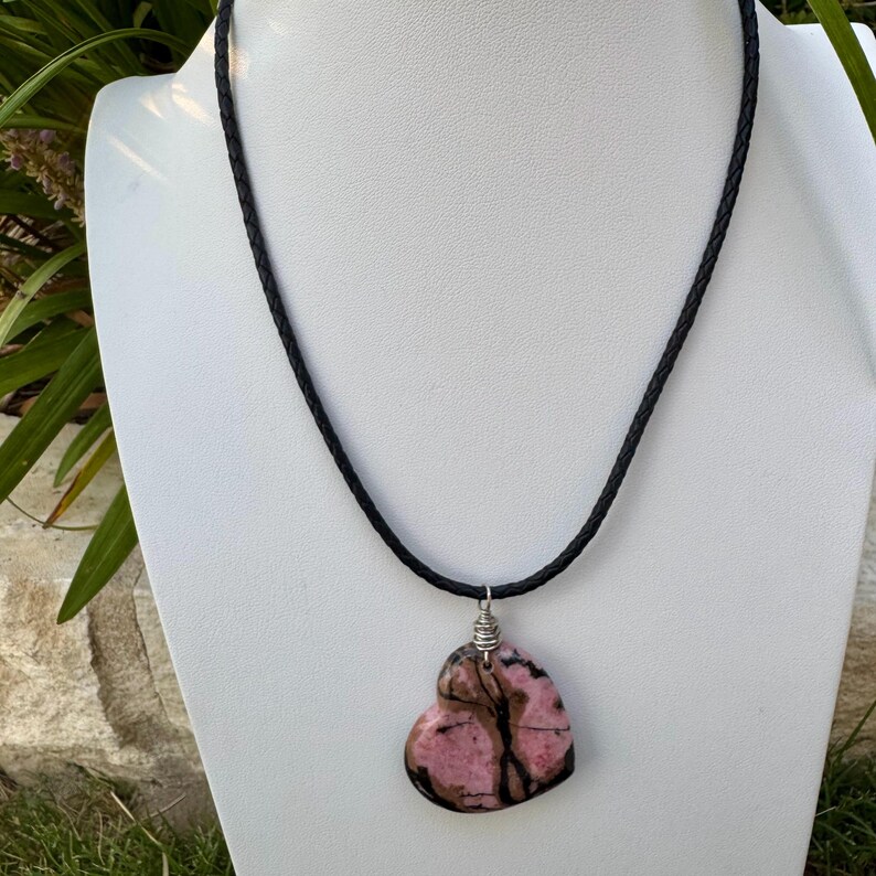 May include: A heart-shaped pendant necklace featuring a pink and brown stone. The pendant is suspended from a black braided cord. The necklace is displayed on a white mannequin.