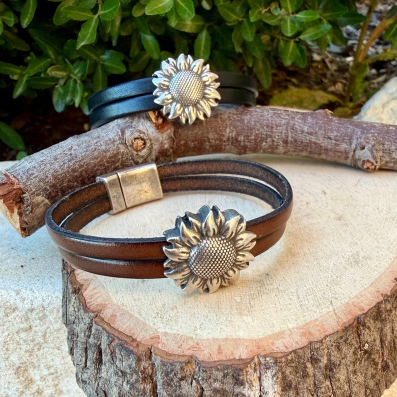 May include: Two leather bracelets with sunflower accents. One bracelet is brown, the other is black. The sunflower is silver-colored. The brown bracelet has a magnetic clasp. The bracelets are on a wooden surface.