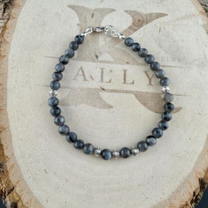 May include: A beaded bracelet featuring round, dark gray labradorite stones interspersed with small, ribbed silver-toned metal spacers. The bracelet is secured with a silver-toned lobster claw clasp and a circular jump ring.
