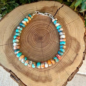 Pode incluir: Uma pulseira de contas de pedra planas e redondas em tons de turquesa, laranja, branco e castanho. As contas est&atilde;o dispostas num padr&atilde;o circular e ligadas por um fecho de mosquet&atilde;o em metal prateado e um pequeno anel de liga&ccedil;&atilde;o.