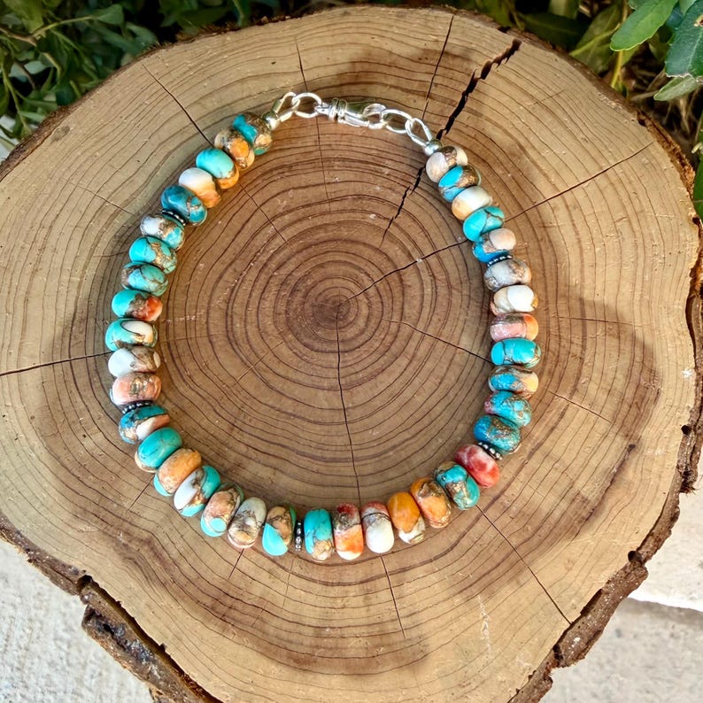 Pode incluir: Uma pulseira de contas de pedra planas e redondas em tons de turquesa, laranja, branco e castanho. As contas est&atilde;o dispostas num padr&atilde;o circular e ligadas por um fecho de mosquet&atilde;o em metal prateado e um pequeno anel de liga&ccedil;&atilde;o.