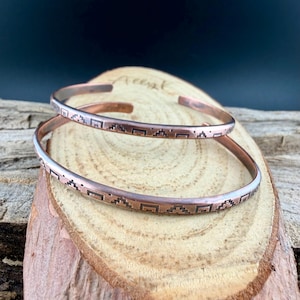 May include: Two copper cuff bracelets featuring a thin, open-ended band design. Each bracelet is engraved with a repeating geometric pattern consisting of small, dark, stepped square motifs that wrap around the exterior surface of the metal.