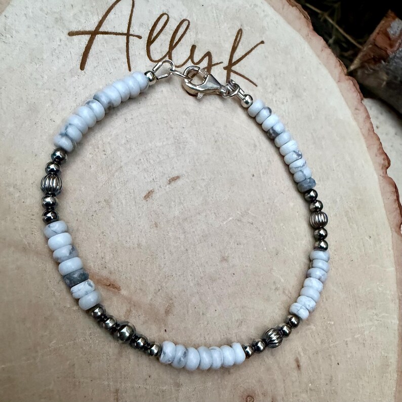 May include: A beaded bracelet featuring white and gray stone beads interspersed with silver-colored metal beads. The bracelet is secured with a silver-colored clasp. The word "Allyk" is written in cursive script in the background.