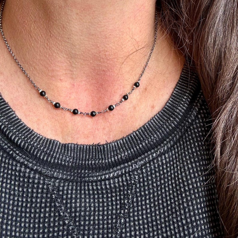 May include: A delicate necklace featuring small, round black beads evenly spaced along a silver-toned chain. The necklace is worn against a dark gray waffle-knit shirt.