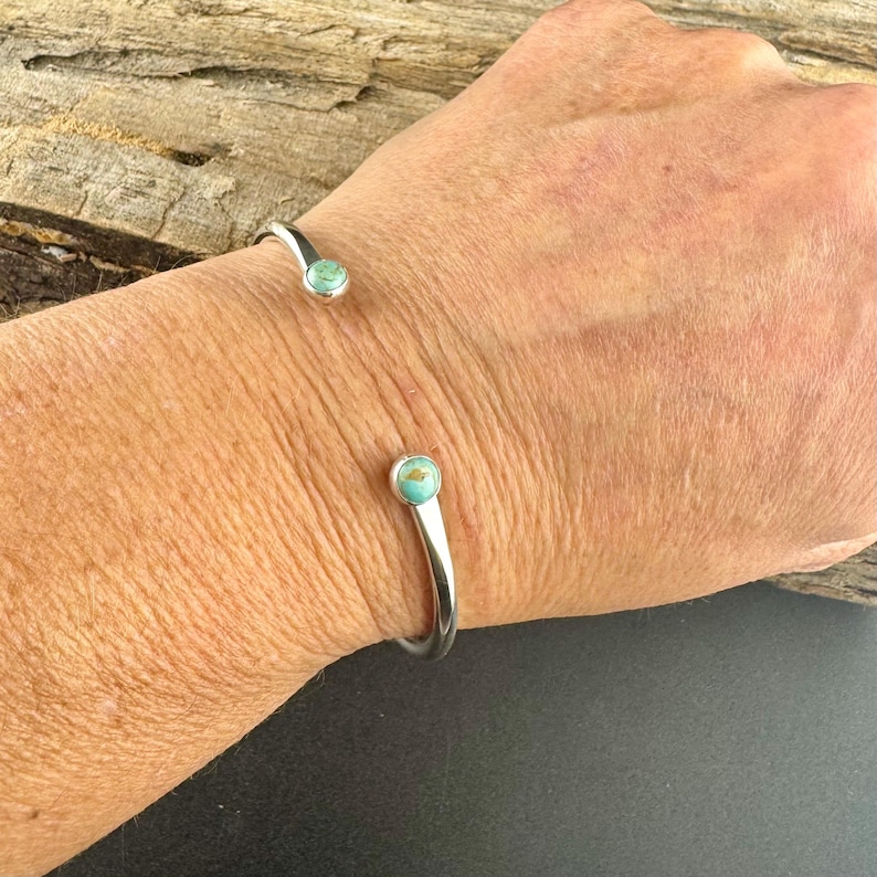K&ouml;nnte beinhalten: Ein silberfarbenes Armreif-Armband mit t&uuml;rkisfarbenen Steinakzenten. Das Armband hat ein schlichtes, offenes Design mit einem abgerundeten Stein an jedem Ende. Das Armband wird am Handgelenk getragen.