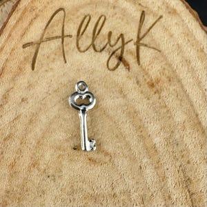 May include: A small, silver-colored key charm with a decorative top and a detailed key bit. The charm is set against a textured, light brown wooden background. The word "Allyk" is written in cursive at the top.