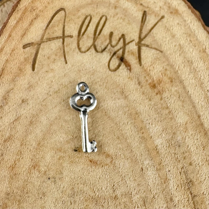 May include: A small, silver-colored key charm with a decorative top and a detailed key bit. The charm is set against a textured, light brown wooden background. The word "Allyk" is written in cursive at the top.