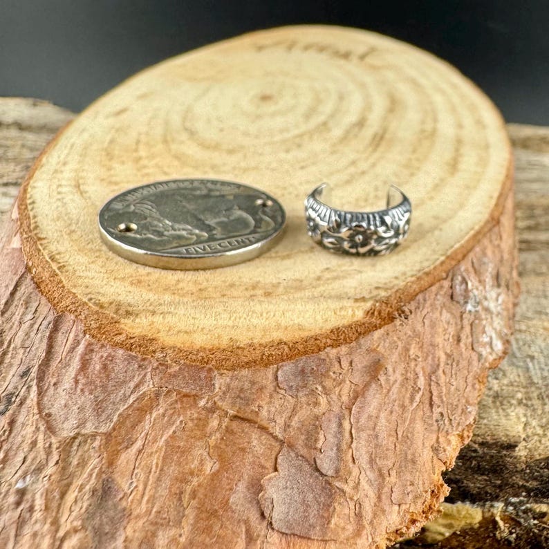 May include: A silver-colored ear cuff with a floral design and a silver coin with a buffalo image. The coin reads "FIVE CENTS". The items are displayed on a wooden surface with a natural wood grain.