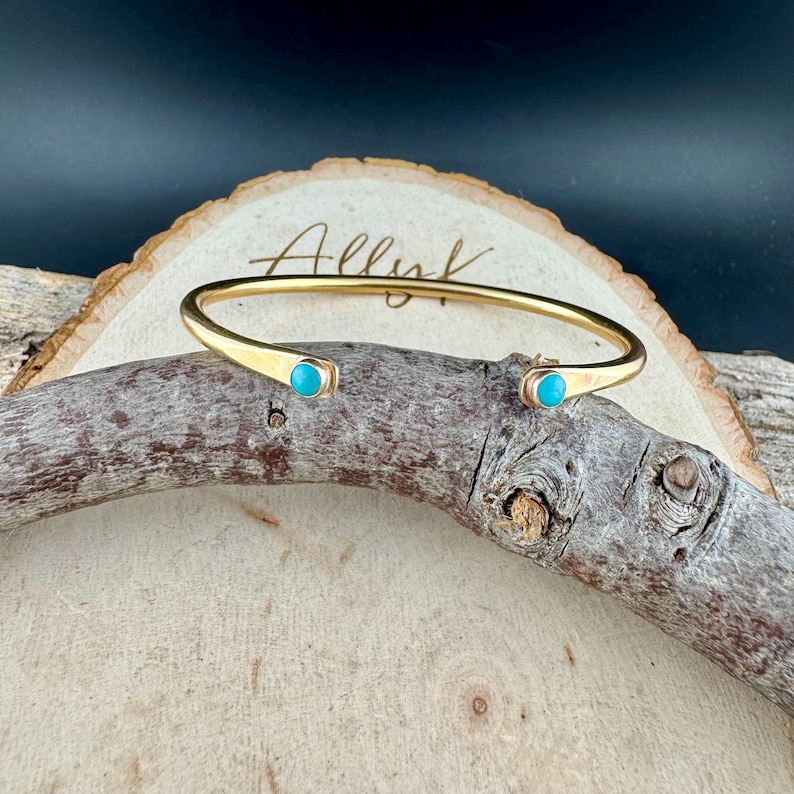 Op de afbeelding: Een goudkleurige manchetarmband met turkooizen stenen aan elk uiteinde. De armband wordt getoond op een stuk hout met het woord "Allure" erop geschreven.