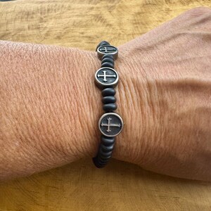 May include: Black beaded bracelet with three silver-toned cross charms. The bracelet is worn on a wrist, showcasing the contrast between the dark beads and the metallic crosses. A simple, yet elegant accessory.