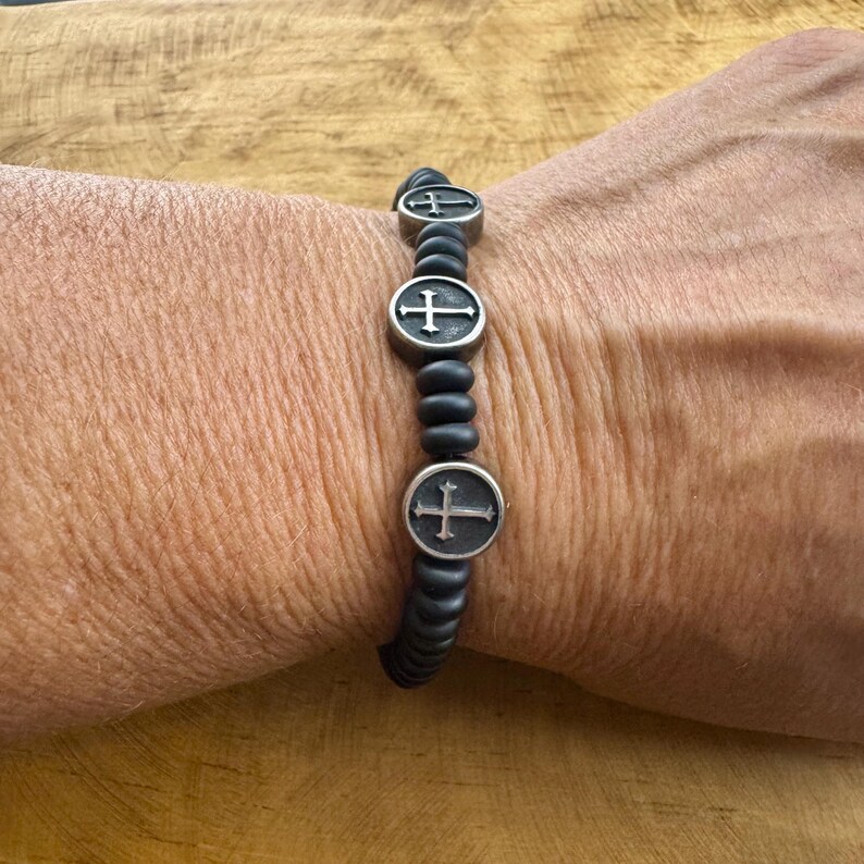 May include: Black beaded bracelet with three silver-toned cross charms. The bracelet is worn on a wrist, showcasing the contrast between the dark beads and the metallic crosses. A simple, yet elegant accessory.