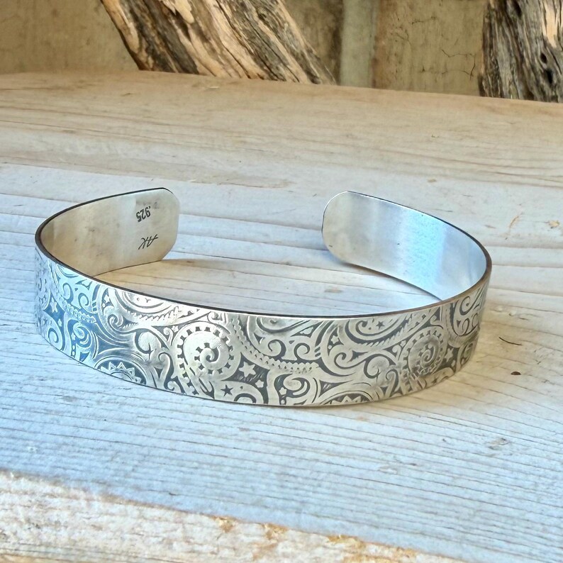 May include: A silver cuff bracelet featuring an intricate, dark-etched pattern of swirling scrolls, spirals, and small stars. The interior of the band is stamped with the text .925 and the initials AK, indicating the sterling silver composition.