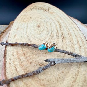 May include: A pair of gold-colored stud earrings with oval, turquoise-colored stones. The earrings are displayed on a wooden surface with a natural, rustic aesthetic. The earrings are set on a wooden branch.