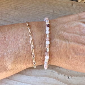 May include: A gold-toned chain bracelet with elongated oval links alongside a beaded bracelet featuring small, smooth, multi-tonal pink and white stone beads interspersed with tiny metallic silver-colored spacers.