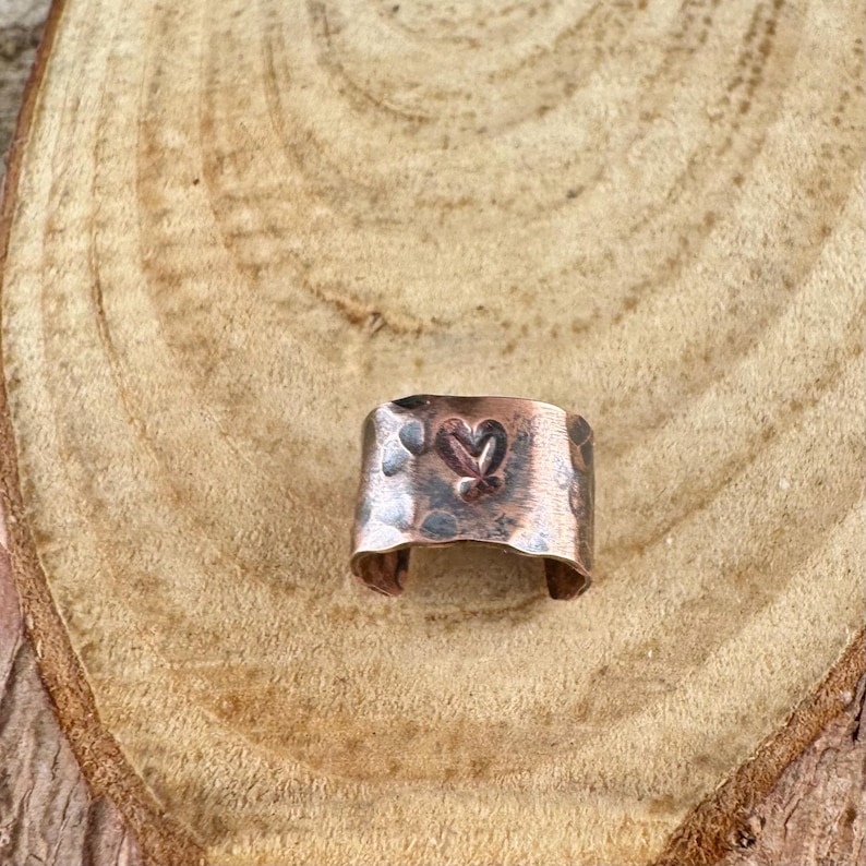 May include: A copper-colored ring with a heart design. The ring has a hammered texture and a cut-out heart in the center. The ring is displayed on a wooden surface.
