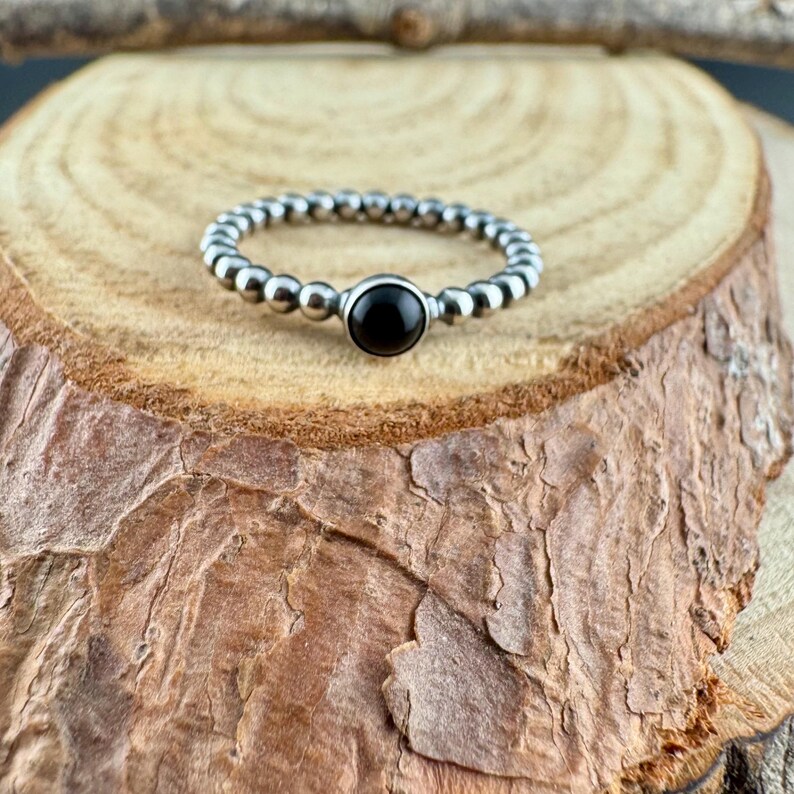 K&ouml;nnte beinhalten: Ein silberner Ring mit einem schwarzen Stein in der Mitte. Das Ringband besteht aus kleinen, runden Perlen. Der Ring wird auf einem St&uuml;ck Holz mit einer nat&uuml;rlichen, strukturierten Oberfl&auml;che pr&auml;sentiert. Der Ring ist ein Schmuckst&uuml;ck.