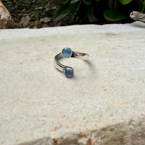 May include: A silver ring with a distinctive open design. The ring has two round, light blue gemstones set at opposing ends of the band. The ring is displayed on a textured, light-coloured surface.