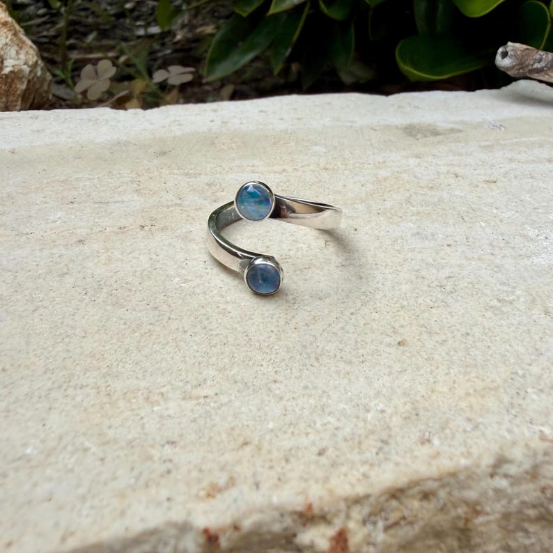May include: A silver ring with a distinctive open design. The ring has two round, light blue gemstones set at opposing ends of the band. The ring is displayed on a textured, light-coloured surface.