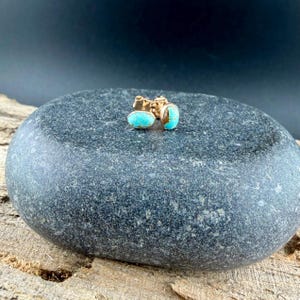 May include: A pair of gold-colored stud earrings with oval turquoise stones. The earrings are displayed on a dark gray, textured stone surface, with a wooden surface in the foreground and a dark background.