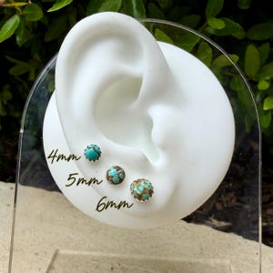 May include: Three turquoise and brown stud earrings, 4mm, 5mm, and 6mm in size, are displayed on a white ear-shaped stand. The earrings have a silver-colored metal setting. The sizes are written in black ink on the clear stand.