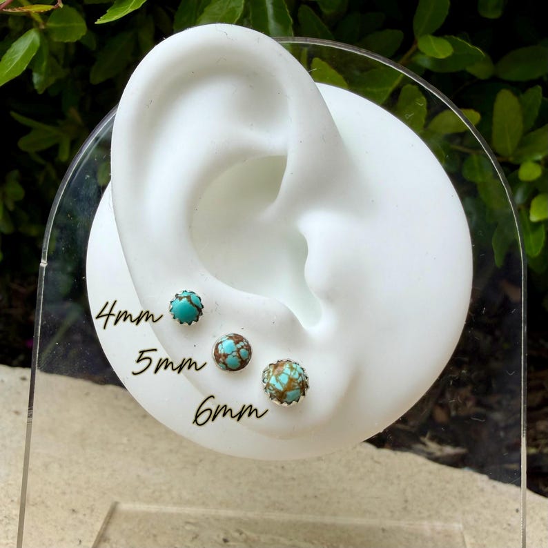 May include: Three turquoise and brown stud earrings, 4mm, 5mm, and 6mm in size, are displayed on a white ear-shaped stand. The earrings have a silver-colored metal setting. The sizes are written in black ink on the clear stand.
