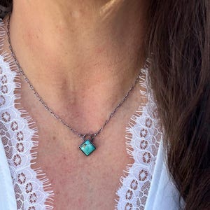 May include: A silver-toned necklace featuring a square turquoise pendant. The pendant is set in a dark metal frame and hangs from a delicate chain. The necklace is worn against a white lace top.