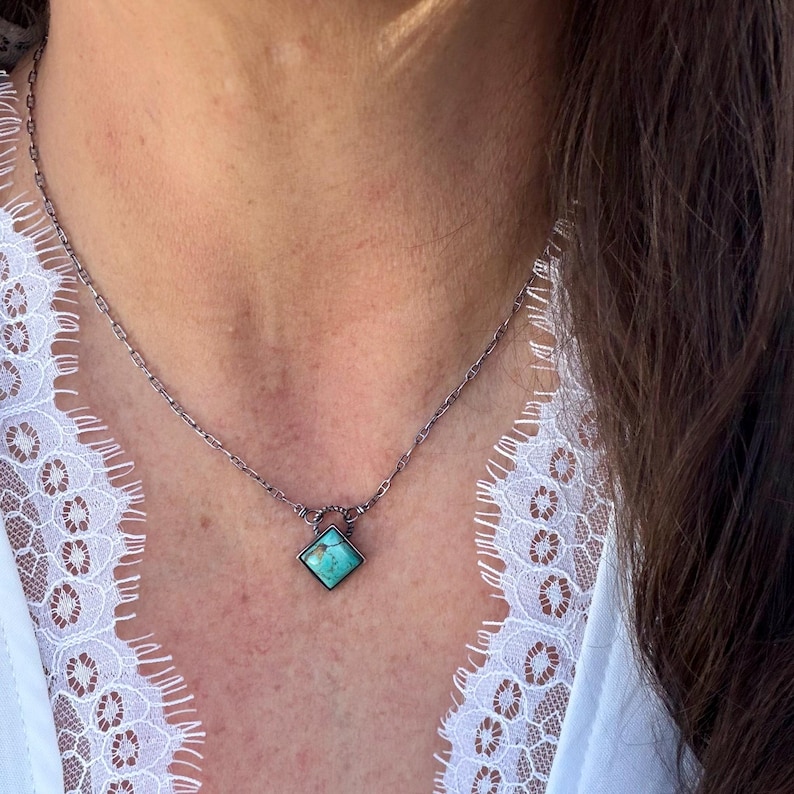 May include: A silver-toned necklace featuring a square turquoise pendant. The pendant is set in a dark metal frame and hangs from a delicate chain. The necklace is worn against a white lace top.