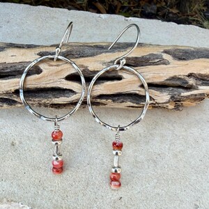 May include: A pair of silver-toned earrings featuring a large, hammered circular hoop suspended from a curved ear wire. Each hoop is accented with a vertical drop consisting of two small, round, reddish-orange beads separated by a silver-toned spacer.