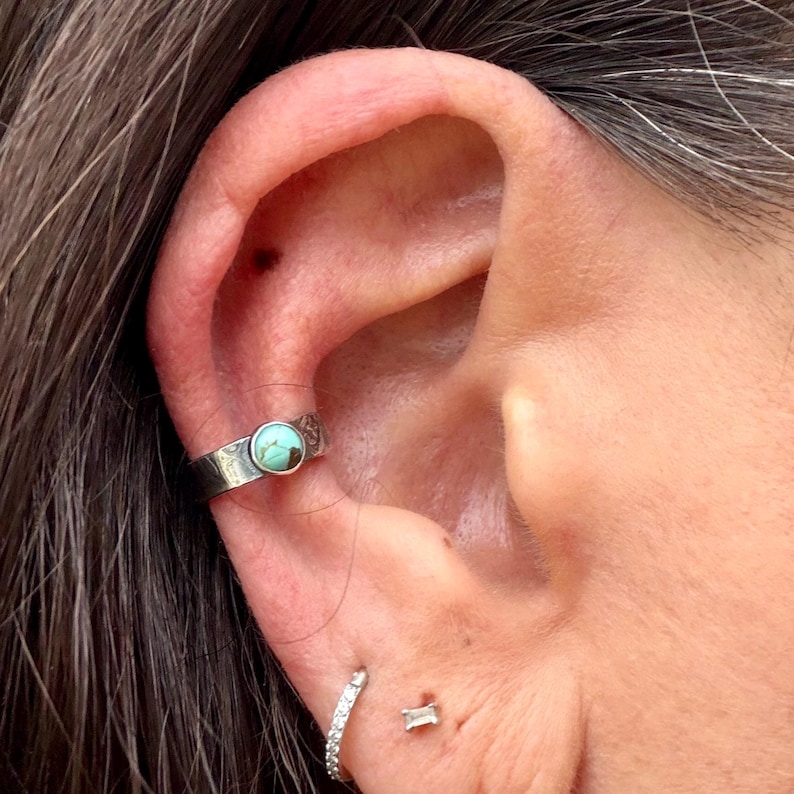 K&ouml;nnte beinhalten: Ein silberner Ohrklemmen-Schmuck mit einem t&uuml;rkisfarbenen Stein. Der Schmuck, der am oberen Ohr getragen wird, hat ein dekoratives Muster. Ein kleiner, diamantbesetzter Ohrring und ein kleiner, rechteckiger Ohrstecker sind ebenfalls sichtbar.