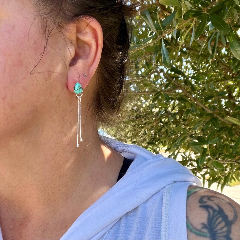 May include: A drop earring featuring a small, teardrop-shaped turquoise stone with dark matrix veining, set in a silver-toned metal mount. Two thin, delicate silver-toned chains hang vertically from the base of the stone, creating a linear design.
