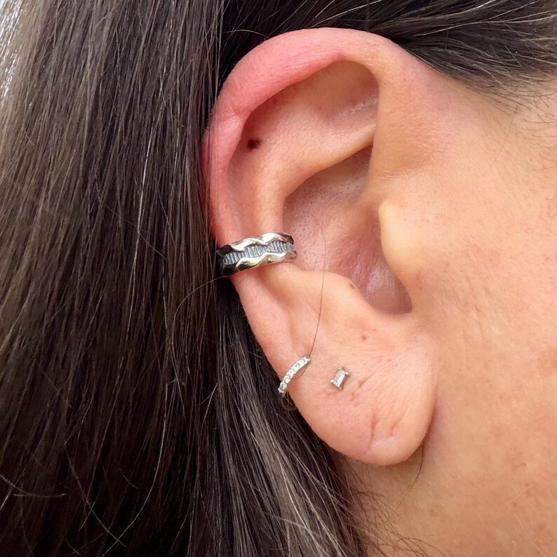 May include: A silver ear cuff with a wavy design is worn on the upper ear. A small, delicate hoop earring with clear stones and a small, rectangular stud earring are also visible. The jewelry is displayed on a person's ear.