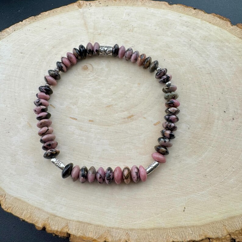 May include: A beaded bracelet featuring pink and black gemstone beads. The bracelet is strung on a silver-colored wire and has silver-colored end caps. The beads are round and vary in size. The bracelet is displayed on a wooden surface.