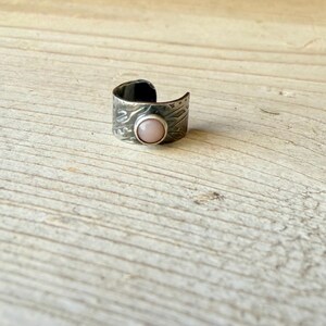May include: An open-band ring crafted from dark, oxidised metal featuring a textured, etched surface pattern and a single, smooth, round, pale pink cabochon stone set securely in the centre of the band's front-facing exterior.