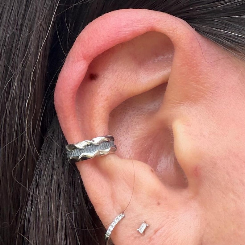 May include: Close-up of an ear adorned with a silver ear cuff featuring a textured, wave-like design. A small, sparkling stud earring and a delicate ring with small clear stones are also visible. The ear has a pinkish hue, and dark hair frames the ear.