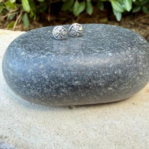 May include: Two small, round, silver-toned stud earrings, each featuring an embossed cross design with four symmetrical arms. The earrings have a polished metallic finish and are displayed as a pair on a smooth, dark gray stone surface.