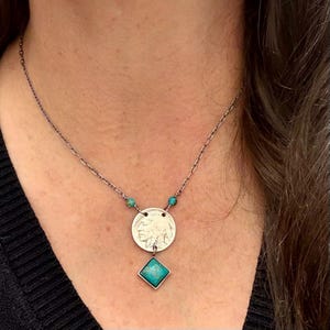 Op de afbeelding: Zilverkleurige ketting met een muntvormige hanger met een Indiaans profiel, geaccentueerd met turkooizen kralen en een vierkante turkooizen stenen hanger. De ketting is delicaat.
