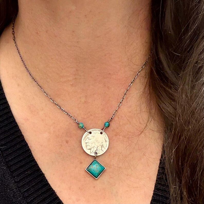 Op de afbeelding: Zilverkleurige ketting met een muntvormige hanger met een Indiaans profiel, geaccentueerd met turkooizen kralen en een vierkante turkooizen stenen hanger. De ketting is delicaat.