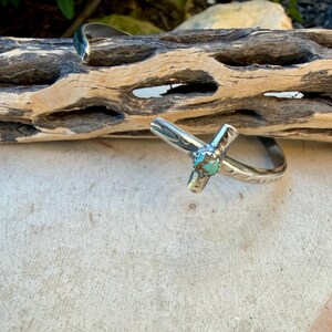May include: A silver cuff bracelet and a silver ring with a cross design. The ring features a turquoise stone at the center of the cross. The jewelry is displayed on a light-colored surface with a wooden background.