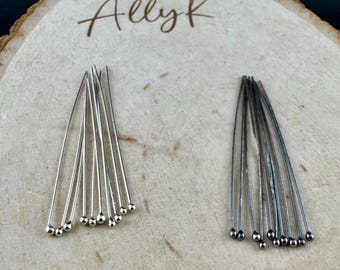 Puede incluir: Dos juegos de alfileres plateados y gris oscuro con pequeños extremos de bola. Los alfileres están dispuestos en forma de abanico sobre una superficie de madera clara. El texto escrito a mano "Allyk" es visible.