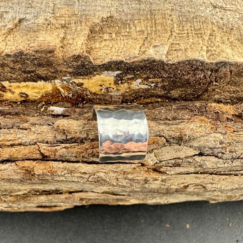 May include: A silver-colored ear cuff with a hammered texture. The cuff has a curved shape and a band of copper-colored metal at the bottom. The background is a piece of wood with a rough, textured surface.