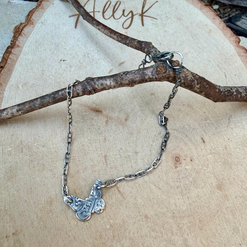 Op de afbeelding: Een zilverkleurige ketting met een vlinderhanger. De vlinderhanger heeft ingewikkelde details. De ketting wordt gepresenteerd op een houten oppervlak met een tak. Het woord "Allyk" is in cursief op het hout geschreven.