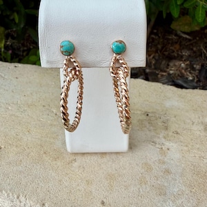 May include: A pair of gold-toned hoop earrings with a twisted rope design. Each earring features a small, round turquoise stone at the top. The earrings are displayed on a white stand against a neutral background.