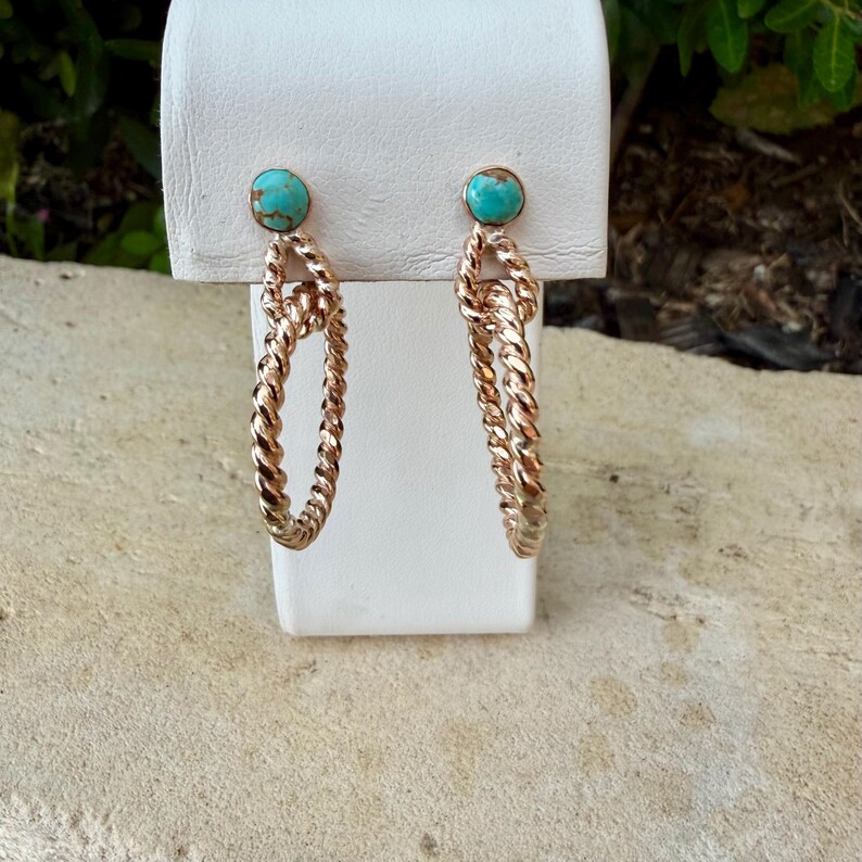 May include: A pair of gold-toned hoop earrings with a twisted rope design. Each earring features a small, round turquoise stone at the top. The earrings are displayed on a white stand against a neutral background.