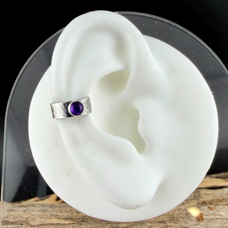 May include: A silver ear cuff with an engraved design and a round, deep purple gemstone. The cuff is displayed on a white ear model, showcasing the jewelry's placement and style. The cuff is a unique accessory.